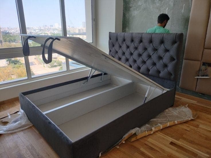 Charcoal grey channel-tufted storage bed with its hydraulic base lifted open, city skyline through the window