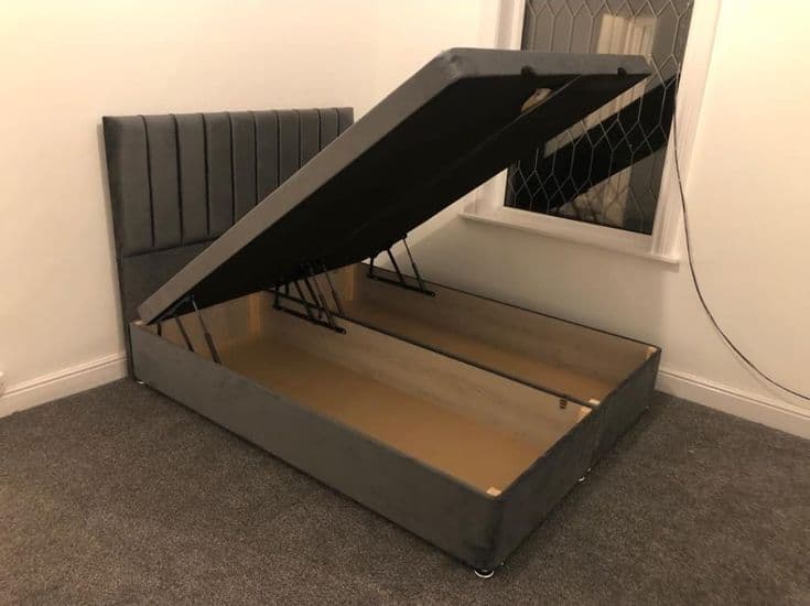 Charcoal grey channel-tufted storage bed with its base lifted open on hydraulic arms, showing the full-width storage beneath
