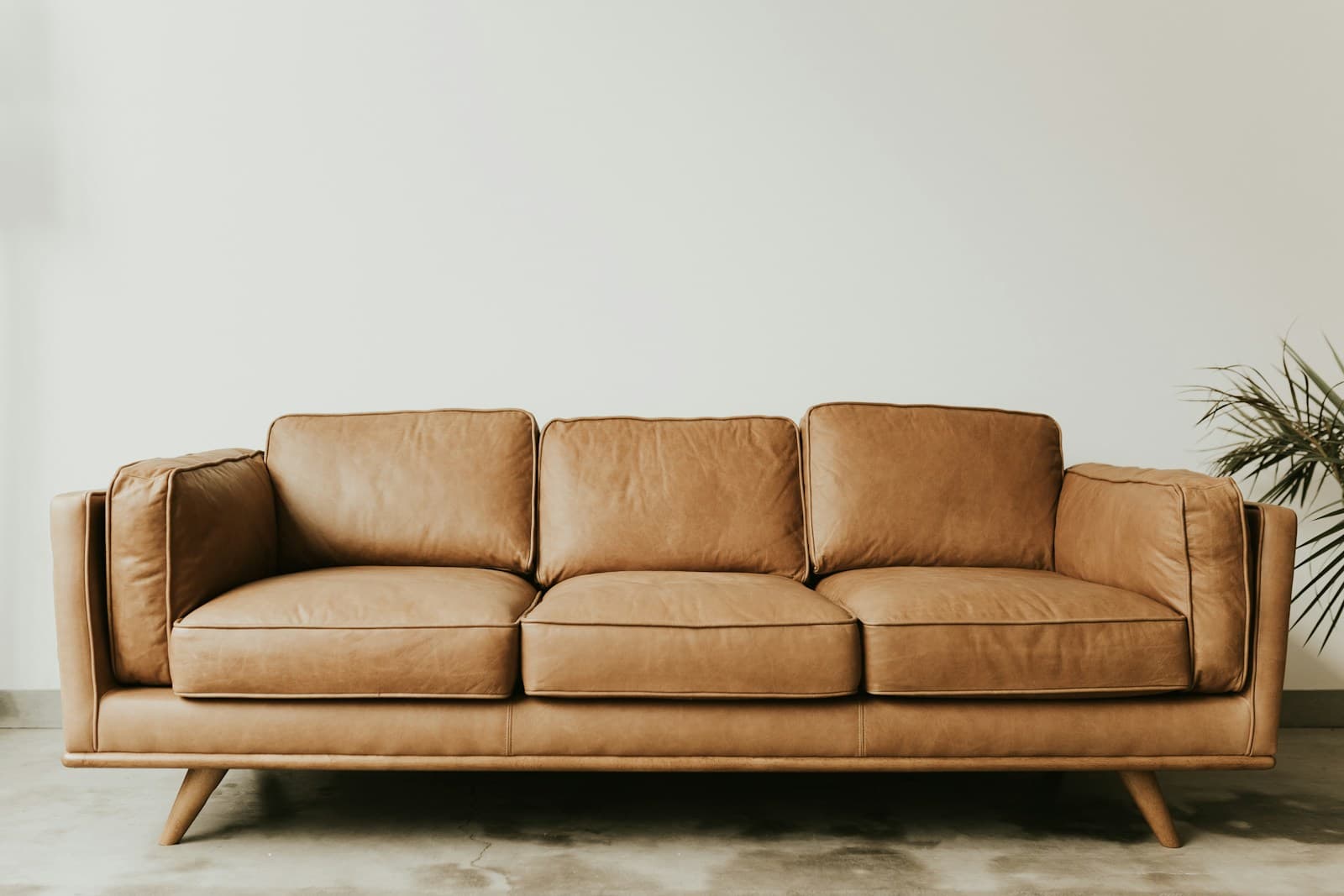 Tan leather three-seater sofa with tapered wooden legs against a pale wall